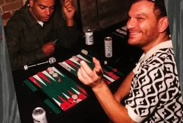 Two players over a backgammon board, drinks on the table